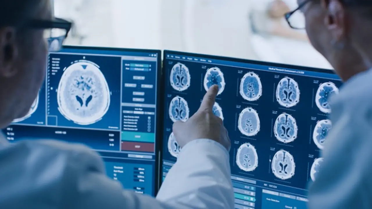 In Control Room Doctor and Radiologist Discuss Diagnosis while Watching Procedure and Monitors Showing Brain Scans Results, In the Background Patient Undergoes MRI or CT Scan Procedure.